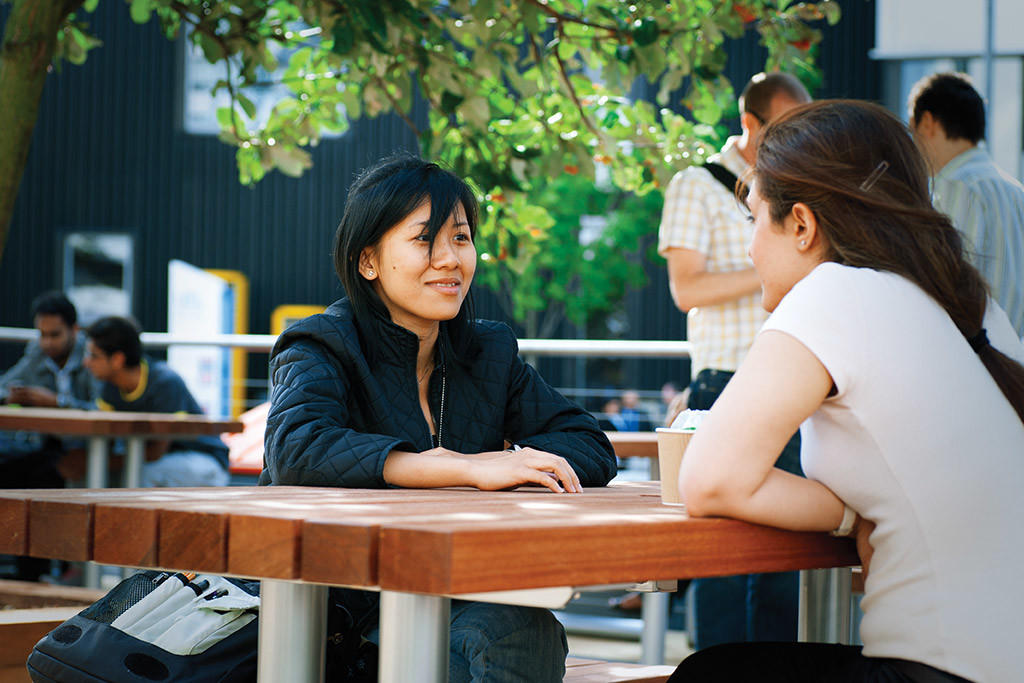 Two women sit at a wooden bench outside