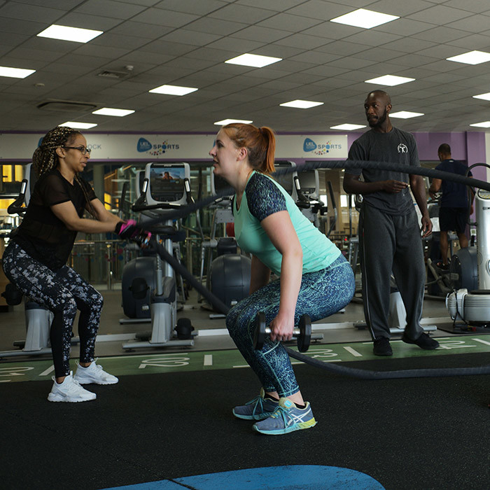 Two women taking part in the circuit class in the gym. One is squatting with weights and one on holding weighted ropes.