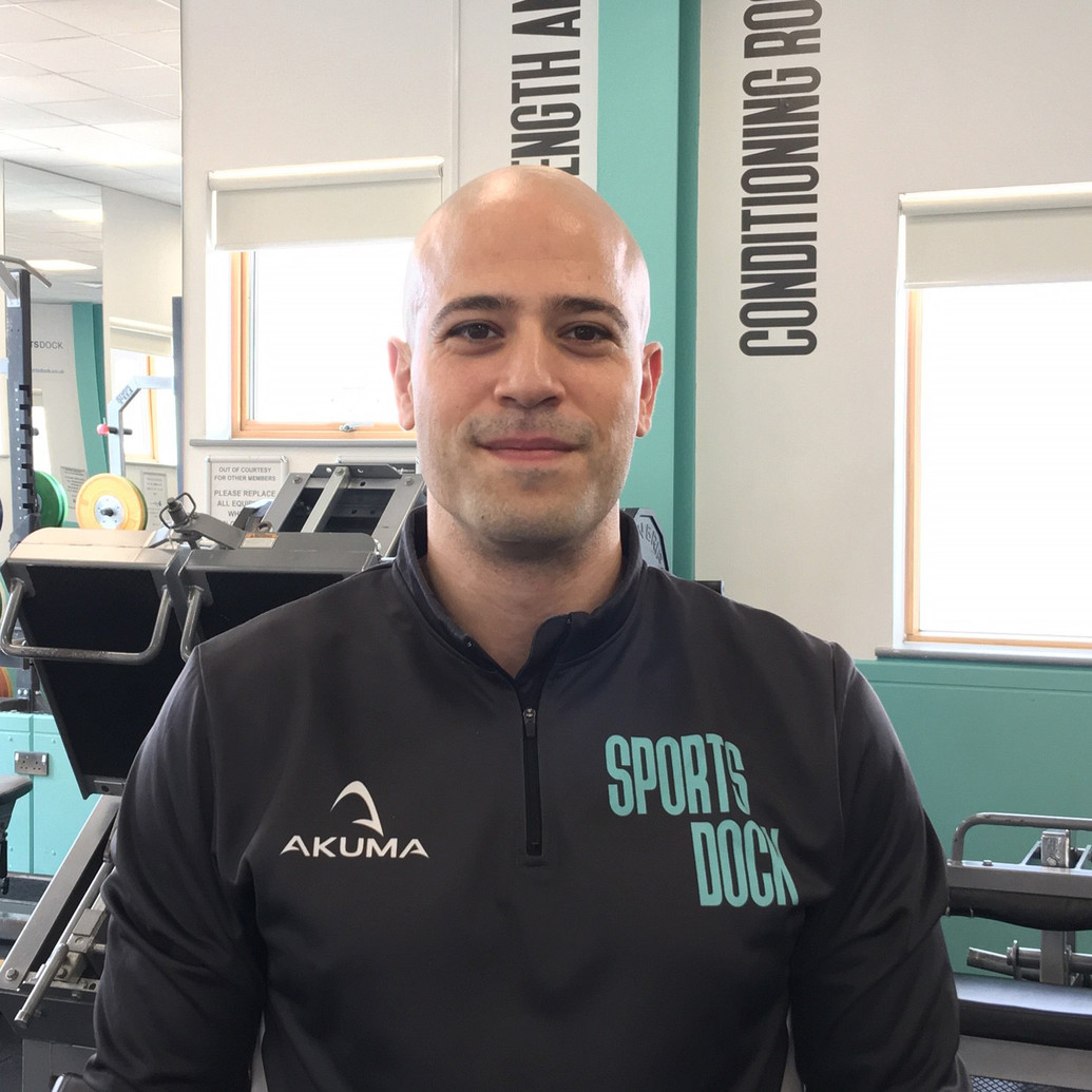 Personal Trainer wearing a black sports jacket with the SportsDock branding stood in the gym in front of a row of treadmills.