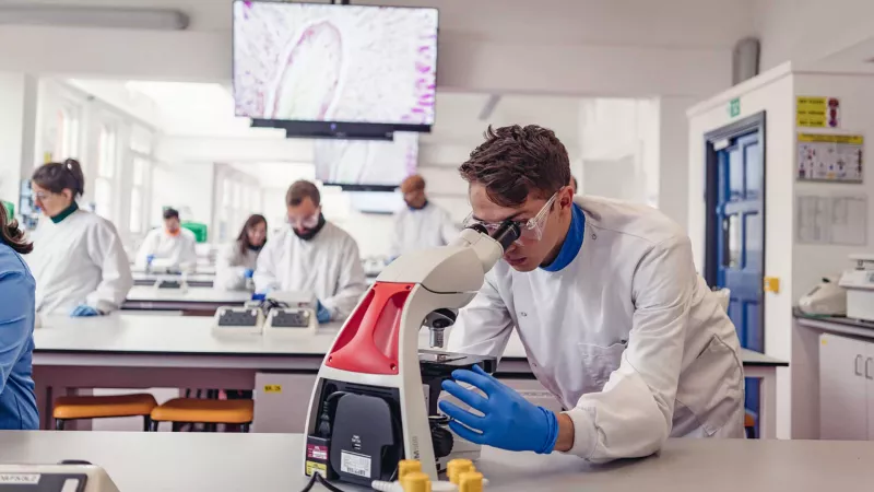Poster of Male student in a white lab coat wearing protective goggles looking into a microscope