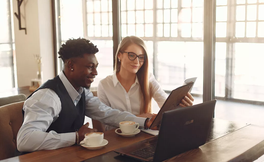 A smiling male and female sitting at a table in front of a laptop and two coffee cups looking at a notebook